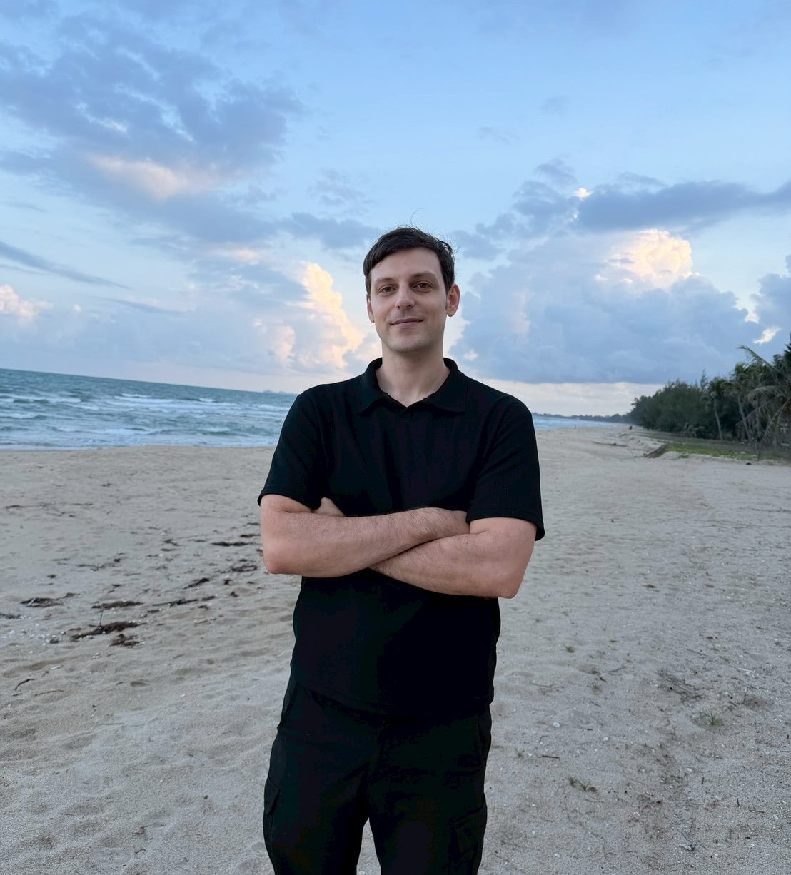 A man standing on a beach with his arms crossed under a blue evening sky.