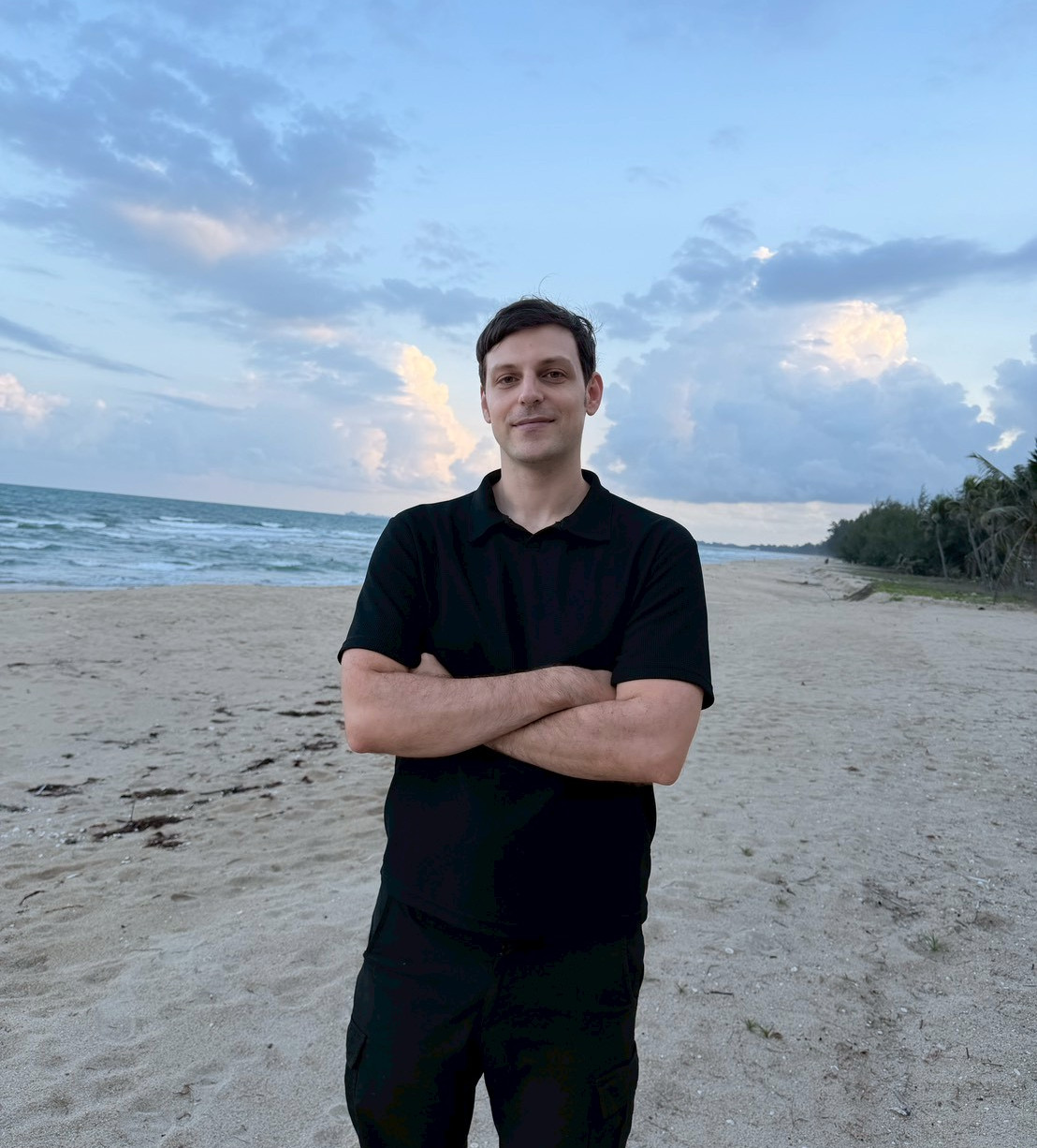 A man standing on a beach with his arms crossed under a blue evening sky.